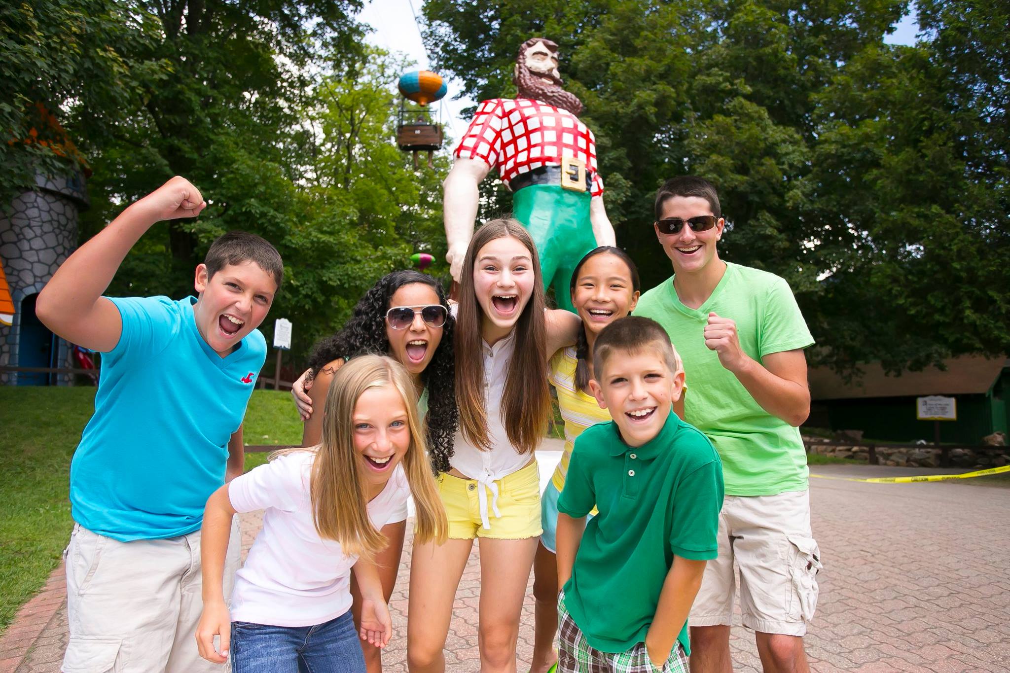 Image of 5 kids standing infant of the Paul Bunyan Statue. The boy on the left is in a green shirt with a huge smile on his face and he is throwing his fist in the air. The little girl inferno of him is leading forward with a grin on her face. The three girls behind her have their arms around each other and smiles as well. Then the boy in the right wearing green is smiling with his fist mid air. Then in the front there is a little boy leaning forward with a smile on his face.